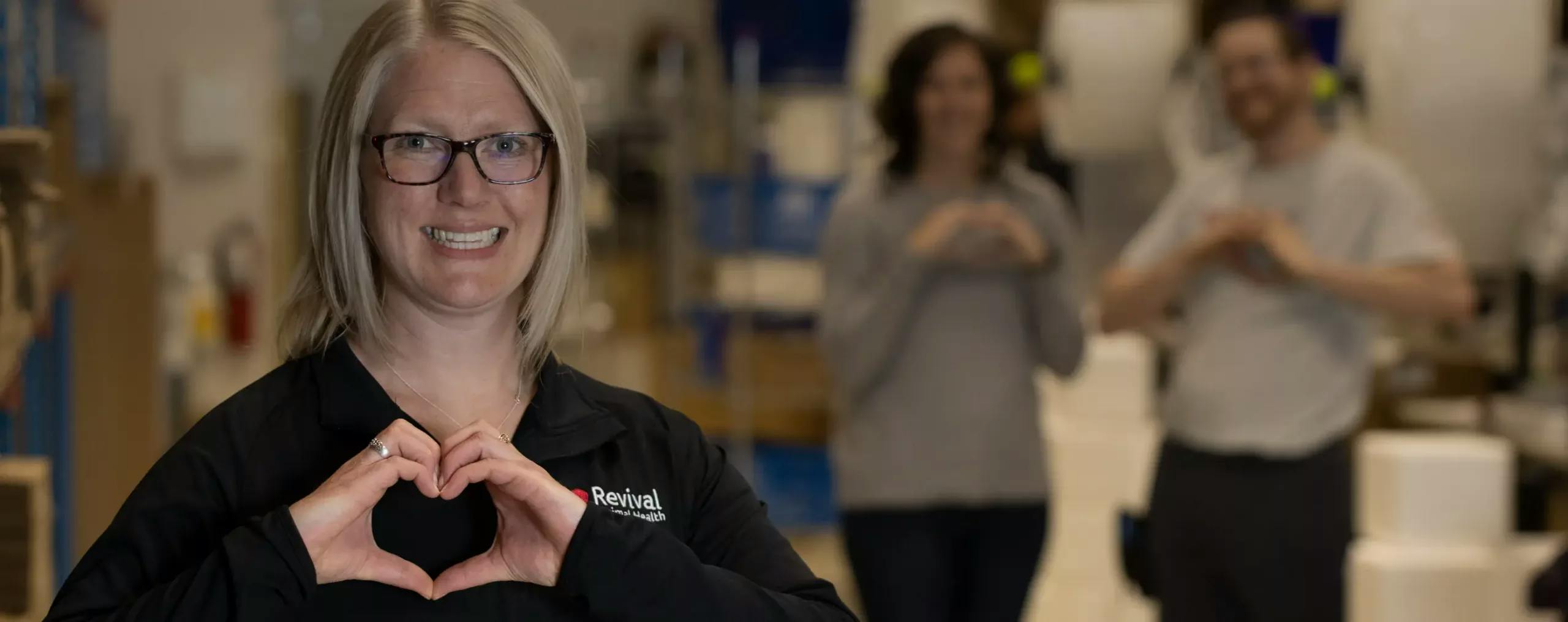 Blonde woman with glasses wearing a black shirt that says Revival makes a heart shape with her hands with a man and a woman behind her in gray shirts are making a heart with their hands.